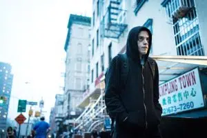 Lebonplanciné - Dans Mr Robot, Rami Malek est Elliot Alderson, un ingénieur en sécurité informatique à Allsafe Security et cyber-justicier qui souffre d’anxiété sociale. Il porte une veste à capuche noire et se déplace dans la rue mains dans les poches.