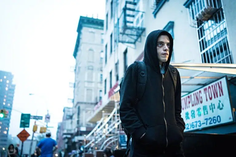Lebonplanciné - Dans Mr Robot, Rami Malek est Elliot Alderson, un ingénieur en sécurité informatique à Allsafe Security et cyber-justicier qui souffre d’anxiété sociale. Il porte une veste à capuche noire et se déplace dans la rue mains dans les poches.