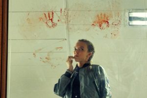 Killing Eve - L'actrice Jodie Comer, dans le rôle de Villanelle, porte un veste une bomber camo et se tient devant un mur couvert de traces de sang faites à la main. Elle reste de marbre.