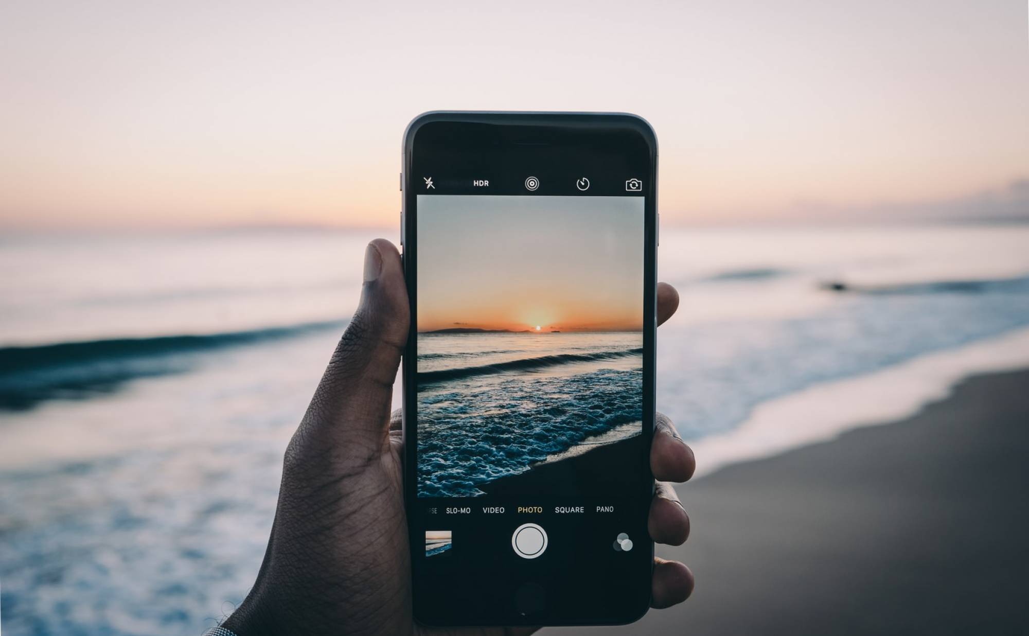 Le bon plan ciné - Main tenant un iPhone et réalisant une vidéo à la plage, au bord de l'eau, sous un couché de soleil